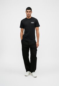 Junger Mann, der ein schwarzes Grafik-T-Shirt, schwarze Cargo-Hosen und weiße Sneakers trägt, vor einem schlichten weißen Hintergrund steht.