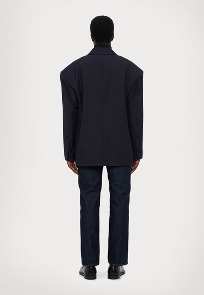 Dunkelblauer, oversized Blazer mit markanten Schulternähten, einem seitlichen Rückenschlitz und passenden Denim-Hosen. Glattes Material und gerades Bein-Design.