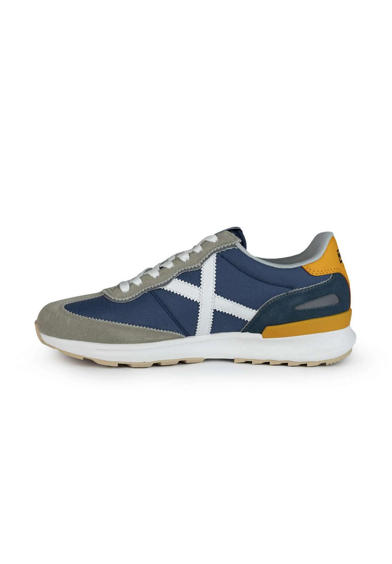 Zapatillas de gamuza y tela en color marino con acentos blancos, parte frontal gris, talón amarillo y suela acolchada. Los cordones son blancos y tienen un diseño de caña baja.