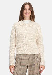 Veste texturée beige avec un col, six boutons et deux poches avant. Portée par-dessus un haut blanc et un pantalon à carreaux.