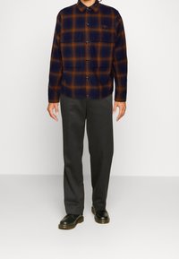 Rutig flanellskjorta i marinblått och orange, med två bröstfickor och svarta knappar, matchad med mörkgrå byxor och svarta skor.