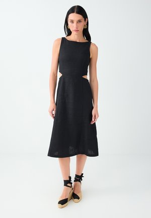 Vrouw met lang donker haar, die een mouwloze zwarte midi-jurk met zijuitsnijdingen en zwarte espadrille sleehakken met enkelbandjes draagt.