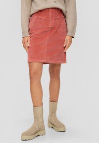 Femme portant une jupe en velours côtelé rose poussiéreux, un pull beige et des bottines en daim beige, se tenant sur un fond gris uni.