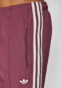 Bordowe spodnie sportowe z gładkiego materiału, z białymi bocznymi paskami, kieszenią na suwak i haftowanym logo w pobliżu krawędzi.