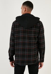 Camicia di flanella a quadri nera con cappuccio nero. Presenta linee bianche e rosse nel motivo. Maniche lunghe con polsini a bottone. Morbido al tatto.
