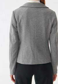 Vrouw in een getailleerde grijze wollen blazer over een witte coltrui, gecombineerd met zwarte broek, afgebeeld van achter tegen een effen achtergrond.