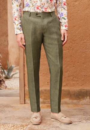 Pantaloni in lino verde oliva dalla vestibilità dritta, con chiusura a bottone, tasche laterali e una texture leggera. Abbinati a mocassini beige.