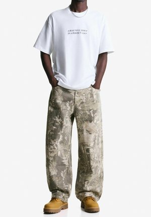 Personne portant une chemise blanche avec le texte "A BEAUTIFUL ERROR IN A PERFECT LOOP", un pantalon cargo ample à fleurs, des bottes jaunes, mains dans les poches.