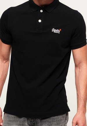 Homme portant un polo noir ajusté avec deux boutons blancs et un petit logo blanc sur le côté gauche de la poitrine, associé à un jean gris.