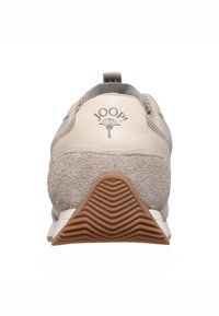 Zapatilla beige con una combinación de cuero suave y gamuza gris. Cuenta con una suela de goma texturizada, un logo en la parte trasera y acentos de malla.