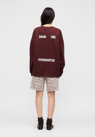 Μπορντό φαρδιά φουτέρ μπλούζα με λευκό κείμενο "BAUM UND PFERDGARTEN" στην πλάτη, συνδυασμένη με σορτς με εκτύπωση λεοπάρ σε μπεζ αποχρώσεις.