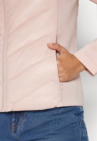 Ljusrosa vadderad jacka med chevronstickning och en slät textur; har en sidoficka och ribbad väv vid kanterna.