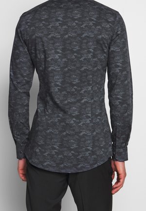 Mężczyzna ubrany w dopasowaną, ciemnoszarą koszulę z długim rękawem o subtelnym pikselowym wzorze kamuflażu, widziany od tyłu.