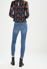 Blouse noire à fleurs avec manches en dentelle transparente, associée à un jean en denim bleu ajusté, présentant un léger effilochage à l'ourlet, et des bottines noires.