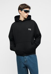 Jeune homme portant un sweat à capuche noir avec poche avant et un jean bleu, regardant sur le côté, et portant des lunettes de soleil noires avec des montures bleues.