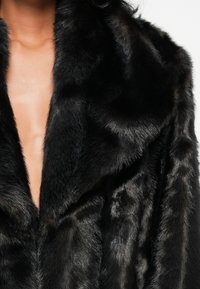 Cappotto in pelliccia sintetica nera con scollatura a V profonda e ampio colletto. Il tessuto presenta una consistenza morbida e liscia e una leggera lucentezza.