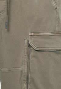 Kaki hlače iz gladkega, trpežnega blaga, z žepom z zadrgo in všitimi poudarki. Vključujejo podrobnost z vrvico blizu pasu.