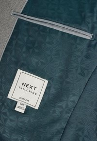 Tissu bleu sarcelle foncé avec un motif géométrique subtil, une petite poche zippée et une étiquette indiquant Next Tailoring Slim Fit 40R fabriqué au Bangladesh.