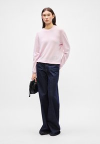 Ροζ πουλόβερ με μακριά μανίκια και στρογγυλή λαιμόκοψη, συνδυασμένο με άνετους σκούρους τζιν με φαρδύ πόδι και μία μαύρη πλεκτή τσάντα.