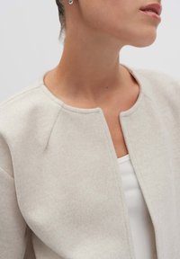 Lichtbeige wollen jacket met een ronde hals, open voorkant en textuurstof. Heeft subtiele stikdetails langs de schouders.