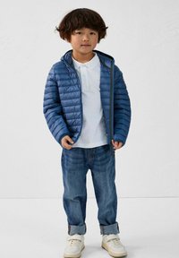 Giacca puffer blu con zip davanti, texture trapuntata, indossata sopra una maglietta polo bianca. Abbinata a jeans blu risvoltati e sneakers bianche.