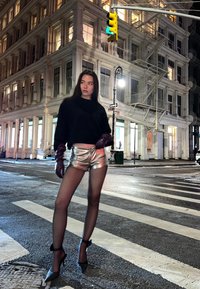Model wearing a black cropped sweater and silver metallic shorts. She has dark sheer tights and black high-heeled shoes with ankle straps.