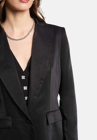 Blazer negro con acabado satinado, solapas estructuradas y dos bolsillos frontales. Debajo, una blusa negra con botones de flor plateados.
