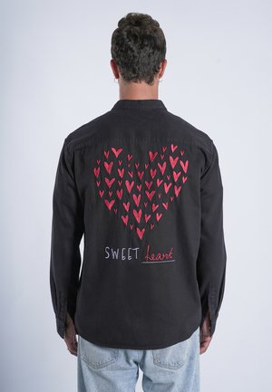 Chemise noire à boutons avec des cœurs rouges brodés formant une forme de cœur dans le dos, accompagnée du texte "SWEET heart" en broderie.