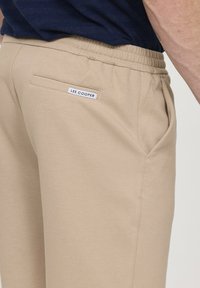 Jogging en beige en coton. Il dispose d'une taille élastiquée, de poches latérales et d'une poche arrière avec une étiquette blanche "Lee Cooper".