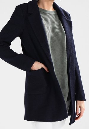 Person trägt einen dunkelmarinefarbenen Mantel über einem grauen, gerippten Pullover, mit einer Hand in der Manteltasche und sichtbarer weißer Hose.