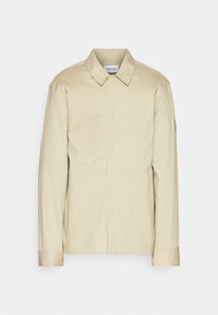 Chaqueta ligera beige con cuello, cierre de botones, mangas largas y un pequeño parche con el logo en la manga. Textura suave.