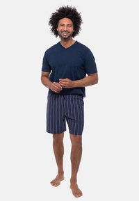 Marineblauwe V-hals t-shirt en gestreepte shorts in blauw, rood en wit. De shorts hebben een elastische tailleband en een lengte tot de knie. Blootsvoets.