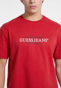 Raudona medvilninė marškinė su apvaliu kaklu. Pasižymi balta ir tamsiai mėlyna spalva teksto: "GUESS JEANS" ir "AMERICAN TRADITION ORIGINAL DESIGNS."