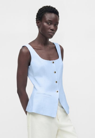 Γυναίκα με σκούρο δέρμα και κοντά σγουρά μαλλιά, που φοράει ανοιχτό μπλε αμάνικο τοπ με κουμπιά και εκρού παντελόνι, στέκεται μπροστά σε απλό φόντο.