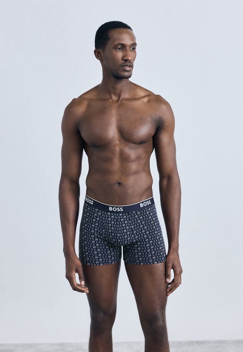 Fekete boxeralsó fehér geometriai mint�ával, rugalmas derékpánttal, melyen fehér "BOSS" logó található. A modell világos háttér előtt pózol.