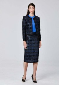 Veste navy sur mesure avec des accents bleus subtils, associée à une jupe courte en tartan navy ajustée. Portée avec un haut bleu vif et des talons noirs.