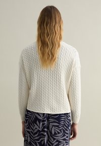 Cremfarbiger Strukturstrickpullover mit verkürztem Design und langen Ärmeln, kombiniert mit einem marineblauen Rock mit weißen Blattmustern.