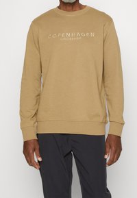 Sweat-shirt beige à manches longues, col rond, avec texte brodé blanc "COPENHAGEN LINDENBERGH" sur le devant. Texture lisse.
