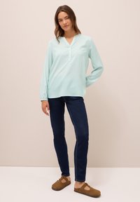 Mujer con blusa de rayas en menta y blanco de manga larga, jeans ajustados oscuros y zapatos cerrados marrones de tipo slip-on, de pie frente a un fondo beige.