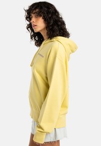 Yellow hoodie in soft cotton, featuring a relaxed fit, long sleeves, and a front pocket. Notable purple logo on the chest.