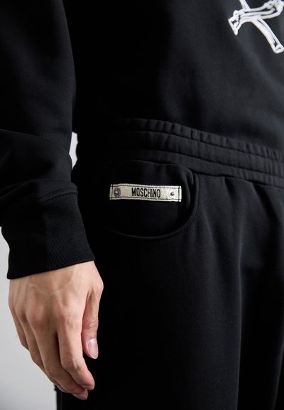 Svarta joggingsbyxor med texturerat tyg, elastisk midja och sidoficka med ett sytt märke som säger "MOSCHINO." Synlig hand.