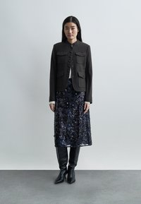 Γυναίκα με μαύρο σακάκι με κουμπιά, μεσαίο φλοράλ δαντελένιο φόρεμα σε ναυτικό μπλε, λευκό εσωτερικό τοπ και μαύρες μπότες που φτάνουν μέχρι το γόνατο, σε απλό φόντο.