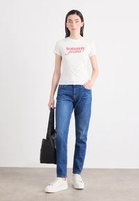 Model nosi belo majico s napisom "tommy jeans" v rdeči barvi, modre kavbojke in bele superge, drži črno torbo na enotnem ozadju.