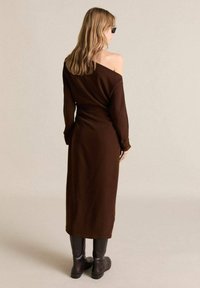 Hnědress off-the-shoulder úpletové šaty s dlouhými rukávy, přiléhavým pasem a dlouhým lemem ke kotníkům. Nosené s černými vysokými botami.
