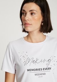 Biały T-shirt z czarnym napisem "Tworzenie Wspomnień Każdego Weekendu", ozdobiony drobnymi metalowymi ćwiekami, okrągłym dekoltem i krótkimi rękawami.