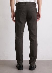 Man wearing dark brown slim-fit pants and black shoes, standing against a plain light wall and floor background, back view.