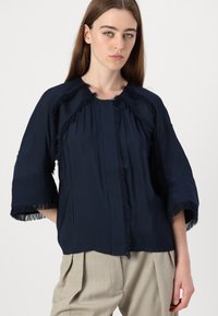 Mørkeblå top med frynsede kanter, korte ærmer og løs pasform. Glat tekstur med en frontplacket og blød drapering.