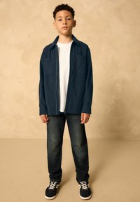 Giacca zipata navy con due tasche frontali indossata su una maglietta bianca, abbinata a jeans scuri e sneakers nere con dettagli bianchi.