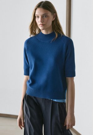 Junge Frau mit lose getragenem langen Haar, die einen blauen kurzärmeligen Strickpullover und schwarze Hosen mit weitem Bein in einem minimalistischen Innenraum trägt.