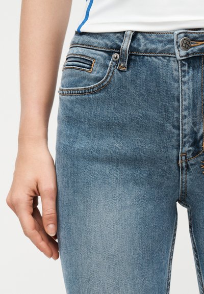 Αριστερό χέρι ακουμπισμένο δίπλα σε ανοιχτό γαλάζιο τζιν με ψηλή μέση, ορατές τσέπες και κουμπωτό φερμουάρ, φορεμένο με λευκό τοπ με μπλε περίγραμμα.